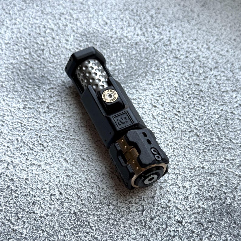 May include: A black and silver metal object with a cylindrical design. The object has a textured silver section and a black casing with gold accents. The text "AMMO CASE" is visible on a small circular emblem.