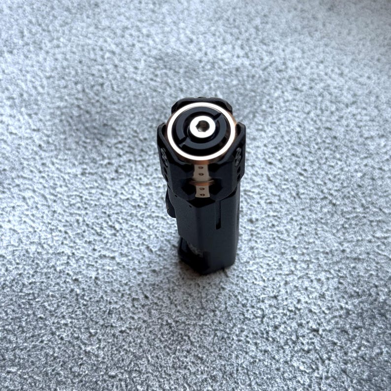 May include: A black electronic device with a circular top featuring a silver ring and a central hole. The device has a rectangular body and is set against a textured gray surface. The top has a gold-colored ring.