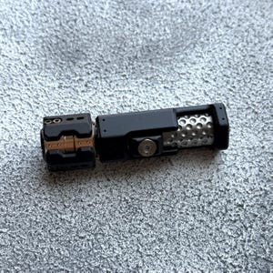 May include: A black metal device with a cylindrical body and a perforated silver section. The device has a rectangular section with a series of holes and a smaller, hinged section with gold-colored accents. The object is on a textured gray surface.
