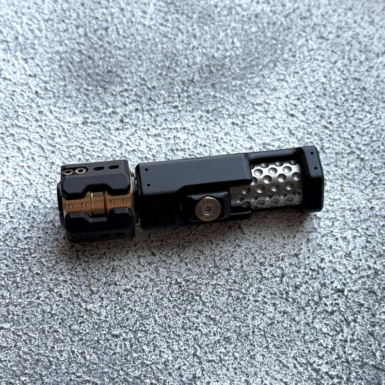 May include: A black metal device with a cylindrical body and a perforated silver section. The device has a rectangular section with a series of holes and a smaller, hinged section with gold-colored accents. The object is on a textured gray surface.