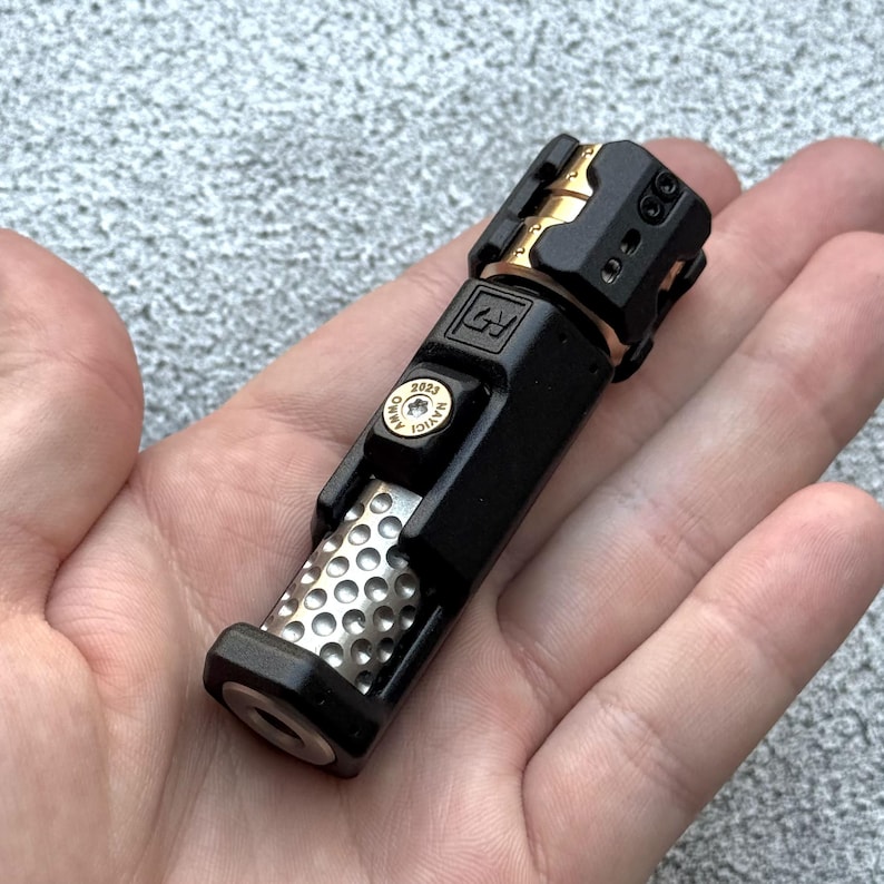 May include: A black and gold metal object with a cylindrical body and a perforated silver section. The object has a complex design with multiple components and the text "2023 AMMO HAYICI" on a gold-colored element. The object is held in a hand.