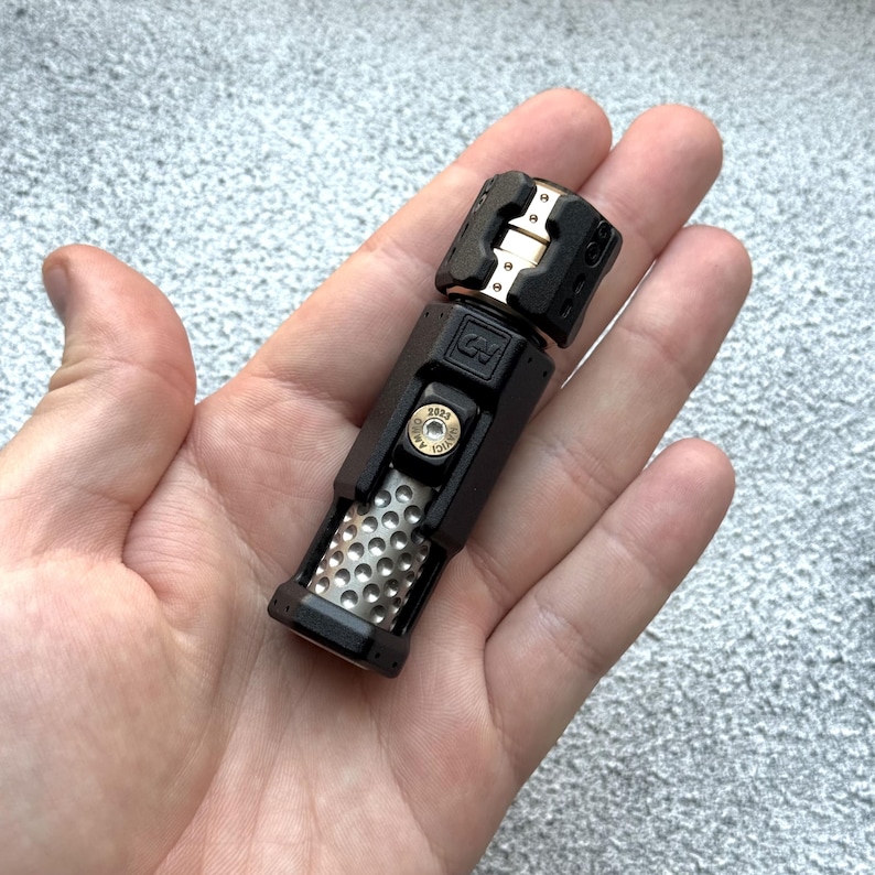 May include: A black and silver metal tool with a complex design. The tool has a cylindrical body with a textured grip and a rectangular top. A gold-colored accent is visible on the top. The tool is held in a hand.