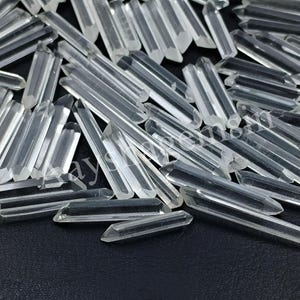 Può includere: Una collezione di punte di cristallo di quarzo trasparente. I cristalli di quarzo sono trasparenti e sfaccettati, con una varietà di forme e dimensioni. I cristalli sono sparsi su una superficie scura.