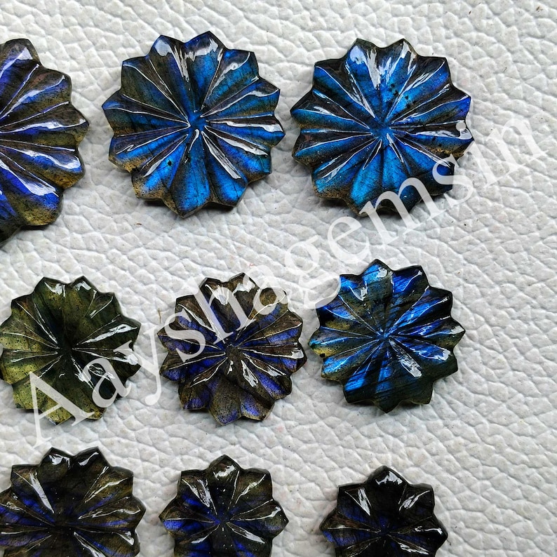 May include: Close-up of several carved labradorite gemstones. The gemstones are starburst-shaped with a blue and green iridescence. The carved details create a textured, dimensional appearance. These gemstones are ideal for jewelry making.