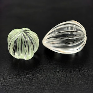 Könnte beinhalten: Zwei dekorative Objekte aus klarem Glas. Eines ist rund mit grünen Akzenten, das andere tropfenförmig. Beide haben ein geripptes Design. Die Objekte werden auf einer schwarzen Oberfläche präsentiert.