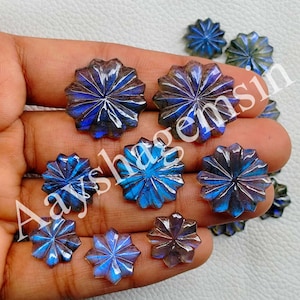 May include: A collection of faceted, starburst-shaped gemstones in shades of blue, purple, and gray. The gemstones are held in a hand, showcasing their intricate carved details and iridescent qualities. The gemstones are likely used for jewelry making.