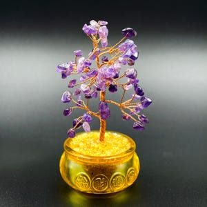 Puede incluir: Un árbol decorativo de piedras preciosas de amatista con un tronco y ramas de alambre de cobre. El árbol está plantado en una pequeña maceta dorada llena de piedras amarillas. Las piedras de amatista son de color púrpura oscuro.