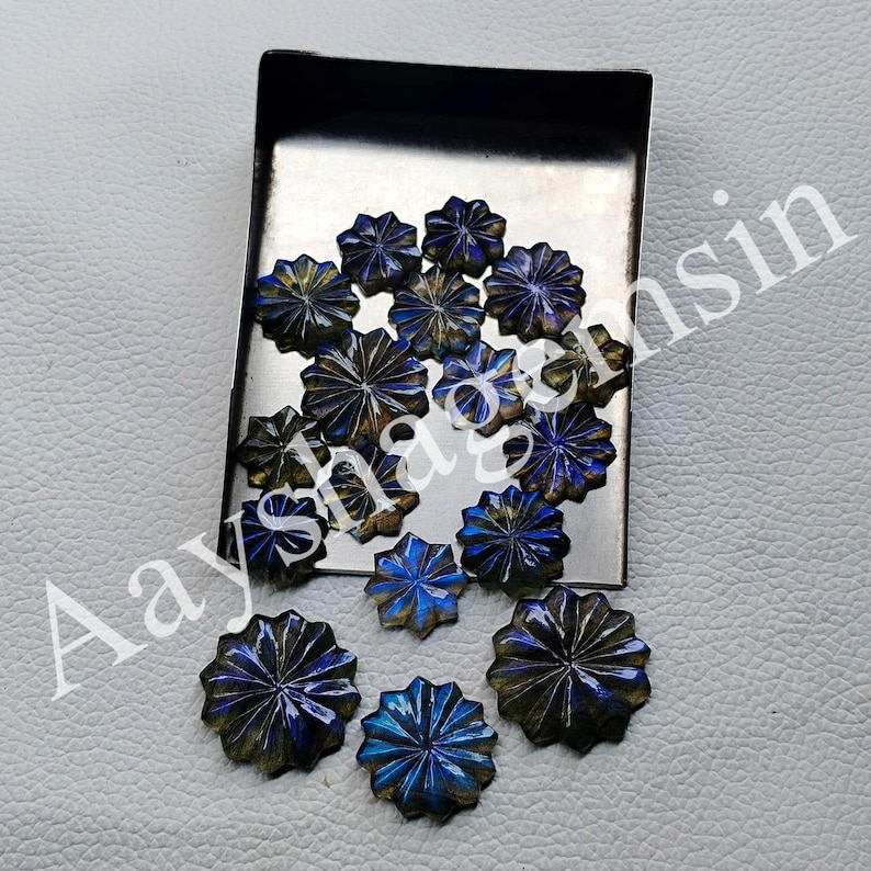 May include: A collection of faceted, starburst-shaped gemstones in shades of blue, gold, and brown. The gemstones are arranged on a small, rectangular metal tray, with some scattered around it. The gemstones have a polished, reflective surface.