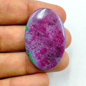 Op de afbeelding: Een ovaalvormige cabochon edelsteen met een levendig magenta en dieproze oppervlak met duidelijke vlekken van heldergroene mineraalinsluitingen en donkere spikkels over het gepolijste, gladde en reflecterende oppervlak van de steen.