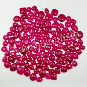 May include: A selection of polished, deep red gemstones in diverse shapes, including squares, ovals, and rounded forms. The gemstones are arranged on a white surface, highlighting their rich colour and smooth texture. The gemstones appear to be cabochons, suitable for jewellery making.