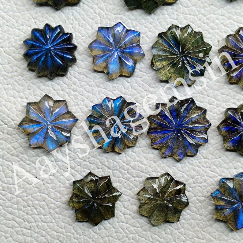 May include: Close-up of several star-shaped labradorite gemstones. The gemstones have a carved design with blue, green, and gold iridescent colors. The gemstones are arranged on a light-colored surface.