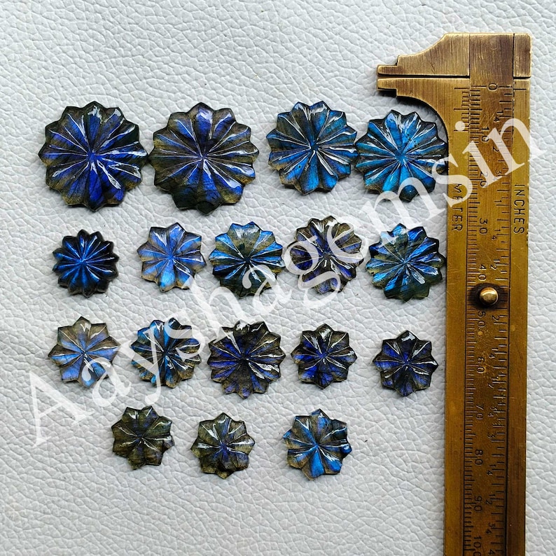 May include: A collection of carved labradorite gemstones in a variety of sizes and shades of blue, gray, and gold. The gemstones are flower-shaped and arranged on a white surface next to a brass ruler.