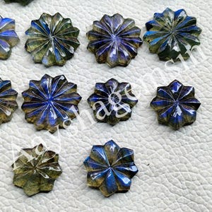 May include: A collection of star-shaped labradorite gemstones. Each gemstone features a carved, multi-faceted design with a mix of blue, green, and gold hues. The gemstones are arranged on a white surface.