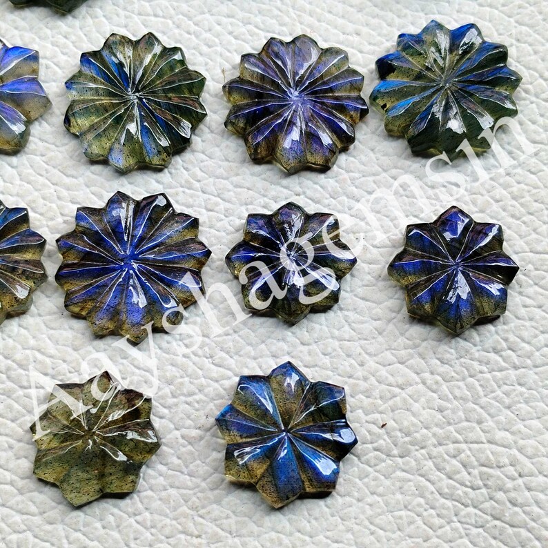 May include: A collection of star-shaped labradorite gemstones. Each gemstone features a carved, multi-faceted design with a mix of blue, green, and gold hues. The gemstones are arranged on a white surface.