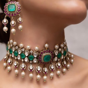 May include: An ornate necklace and matching earrings set. The jewelry features emerald green and ruby red gemstones, surrounded by clear crystals and dangling pearls. The metal has a gold tone. The earrings hang from the ear, and the necklace sits around the neck.