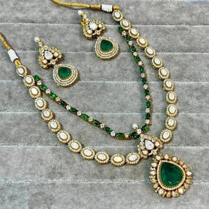 K&ouml;nnte beinhalten: Ein goldfarbenes Halsketten- und Ohrring-Set mit smaragdgr&uuml;nen und klaren Edelsteinen. Die Halskette hat ein mehrschichtiges Design mit einem gro&szlig;en tropfenf&ouml;rmigen Anh&auml;nger. Die Ohrringe haben ein passendes Design.