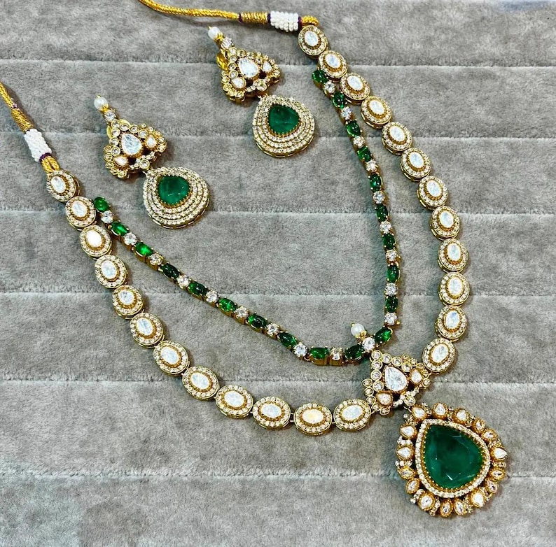 K&ouml;nnte beinhalten: Ein goldfarbenes Halsketten- und Ohrring-Set mit smaragdgr&uuml;nen und klaren Edelsteinen. Die Halskette hat ein mehrschichtiges Design mit einem gro&szlig;en tropfenf&ouml;rmigen Anh&auml;nger. Die Ohrringe haben ein passendes Design.