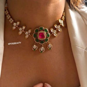 May include: A gold-toned choker necklace featuring a central floral-shaped pendant with a square pink stone surrounded by green stones. The chain consists of repeating leaf-shaped elements with clear stones and three dangling teardrop charms. Text reads XPOSEBEAUTY.