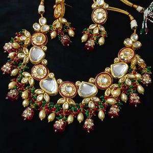 Puede incluir: Un conjunto de collar y pendientes dorados sobre fondo negro. El collar presenta piedras transparentes en forma de l&aacute;grima, cuentas rojas y verdes y perlas de imitaci&oacute;n. Los pendientes reflejan el dise&ntilde;o del collar. Joyas para ocasiones especiales.