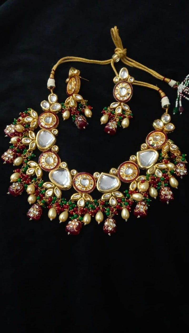 Puede incluir: Un conjunto de collar y pendientes dorados sobre fondo negro. El collar presenta piedras transparentes en forma de l&aacute;grima, cuentas rojas y verdes y perlas de imitaci&oacute;n. Los pendientes reflejan el dise&ntilde;o del collar. Joyas para ocasiones especiales.