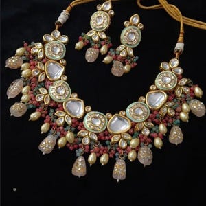 Puede incluir: Un collar y un juego de pendientes a juego en tono dorado. El collar presenta piedras grandes, claras y facetadas, rodeadas de filigrana dorada y cuentas m&aacute;s peque&ntilde;as y coloridas. Los pendientes reflejan el dise&ntilde;o del collar. El conjunto est&aacute; sobre un fondo negro.