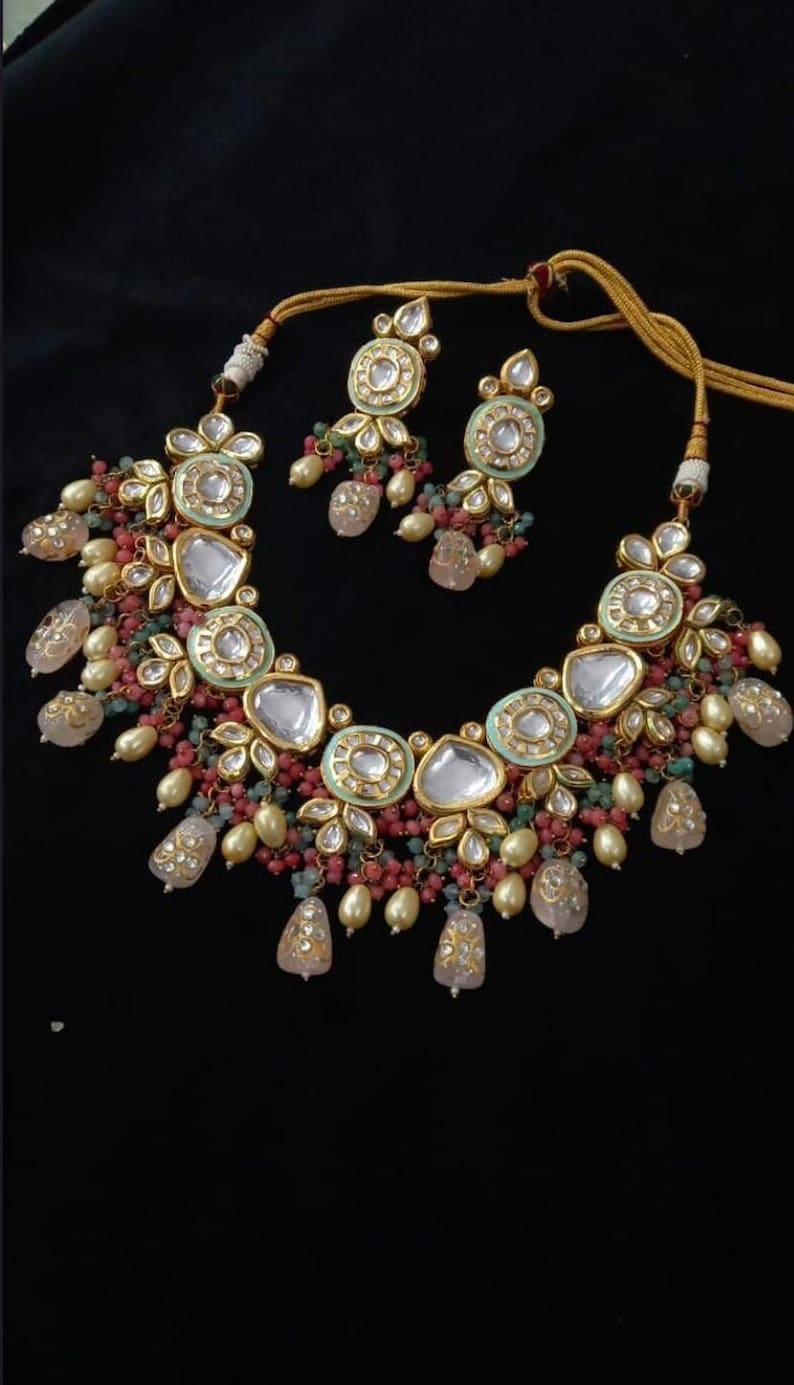 Puede incluir: Un collar y un juego de pendientes a juego en tono dorado. El collar presenta piedras grandes, claras y facetadas, rodeadas de filigrana dorada y cuentas m&aacute;s peque&ntilde;as y coloridas. Los pendientes reflejan el dise&ntilde;o del collar. El conjunto est&aacute; sobre un fondo negro.