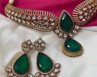 Op de afbeelding: Een goudkleurige sieradenset met een ketting en oorbellen. De ketting en oorbellen hebben grote, druppelvormige groene edelstenen. De ketting en oorbellen zijn versierd met kleine, heldere edelstenen.