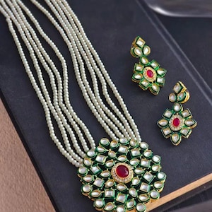 Puede incluir: Un collar de perlas y un conjunto de pendientes a juego. El collar presenta múltiples hilos de pequeñas perlas blancas. El colgante y los pendientes tienen un diseño verde y dorado con una piedra roja central y piedras blancas.