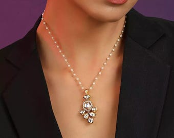 Puede incluir: Un collar con colgante dorado con un grupo de piedras claras en forma de lágrima y una piedra central rectangular con un halo de piedras más pequeñas. El collar tiene una delicada cadena de pequeñas perlas blancas. La persona lleva una blazer negro.