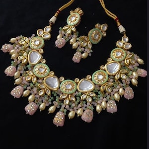Puede incluir: Un conjunto de collar y pendientes a juego en tono dorado. El collar presenta una serie de colgantes circulares de color verde claro y dorado con detalles de piedras transparentes. Cuentas rosas y crema y colgantes en forma de l&aacute;grima cuelgan del collar y los pendientes.