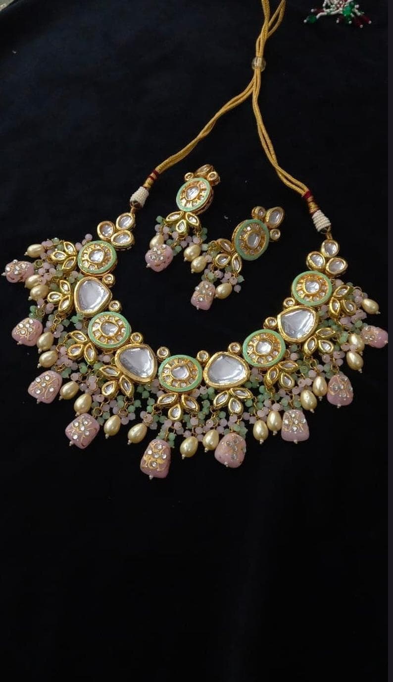 Puede incluir: Un conjunto de collar y pendientes a juego en tono dorado. El collar presenta una serie de colgantes circulares de color verde claro y dorado con detalles de piedras transparentes. Cuentas rosas y crema y colgantes en forma de l&aacute;grima cuelgan del collar y los pendientes.
