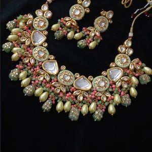 Puede incluir: Un conjunto de collar y pendientes en tono dorado con una combinaci&oacute;n de colores verde menta, rosa y crema. El collar presenta piedras grandes y transparentes en forma de l&aacute;grima y piedras redondas m&aacute;s peque&ntilde;as. Los pendientes reflejan el dise&ntilde;o del collar.