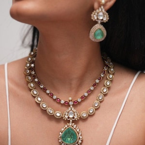 Pode incluir: Um conjunto de colar e brincos dourados com pedras preciosas verdes esmeralda e transparentes. O colar tem um grande pingente em forma de gota com uma pedra verde. Os brincos têm um design semelhante. A joia é cravejada com pedras pequenas e transparentes.