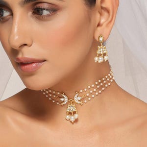 May include: Gold-coloured bridal jewellery set featuring a layered pearl necklace and matching earrings. The necklace has a floral pendant with dangling pearls and a crescent moon design. The earrings have a similar design with dangling pearls.