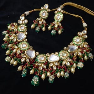 Puede incluir: Un conjunto de collar y pendientes en tono dorado con detalles blancos, verdes y rojos. El collar presenta piedras grandes y claras en forma de l&aacute;grima y piedras redondas m&aacute;s peque&ntilde;as. Los pendientes reflejan el dise&ntilde;o del collar. Sobre fondo negro.