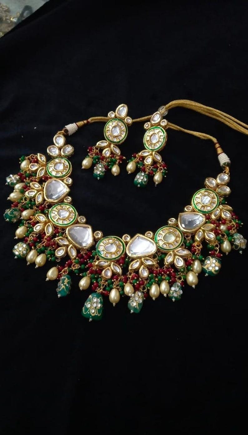 Puede incluir: Un conjunto de collar y pendientes en tono dorado con detalles blancos, verdes y rojos. El collar presenta piedras grandes y claras en forma de l&aacute;grima y piedras redondas m&aacute;s peque&ntilde;as. Los pendientes reflejan el dise&ntilde;o del collar. Sobre fondo negro.