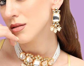 Puede incluir: Un conjunto de collar y pendientes dorados con piedras claras, cuadradas y en forma de lágrima. El collar tiene un hilo de cuentas blancas y los pendientes tienen una piedra cuadrada con perlas colgantes. La joyería está sobre un fondo morado.