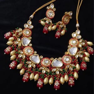 Puede incluir: Un conjunto de collar y pendientes en tono dorado. El collar presenta detalles rojos, verdes y dorados con incrustaciones de piedras blancas. Los pendientes reflejan el dise&ntilde;o del collar, creando un conjunto de joyer&iacute;a coordinado.