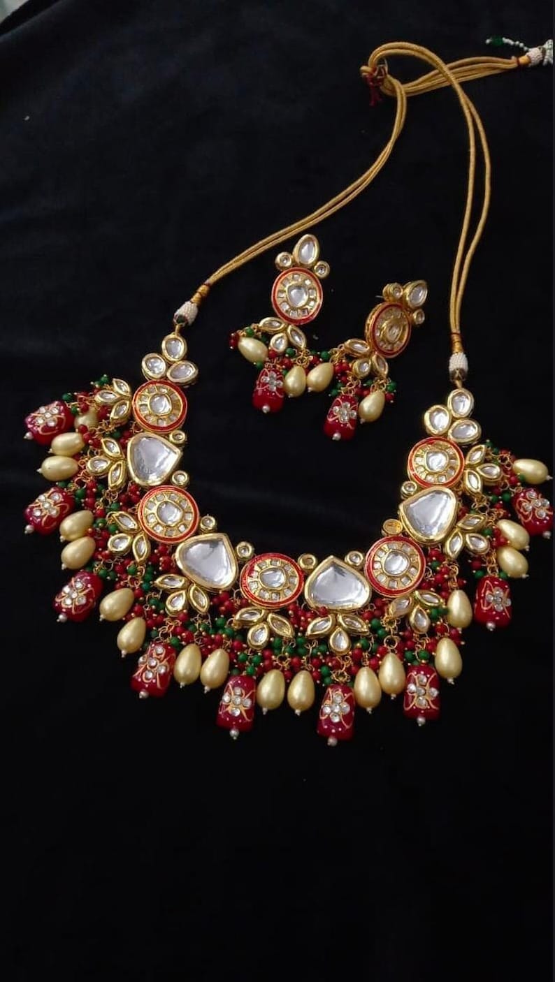 Puede incluir: Un conjunto de collar y pendientes en tono dorado. El collar presenta detalles rojos, verdes y dorados con incrustaciones de piedras blancas. Los pendientes reflejan el dise&ntilde;o del collar, creando un conjunto de joyer&iacute;a coordinado.