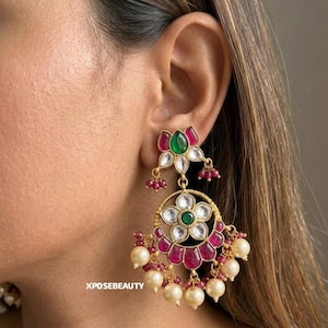 Puede incluir: Pendientes dorados con diseño floral. Los pendientes presentan una parte superior en forma de flor con piedras verdes y rosas, y un colgante circular con piedras blancas, rosas y verdes, y perlas colgantes.