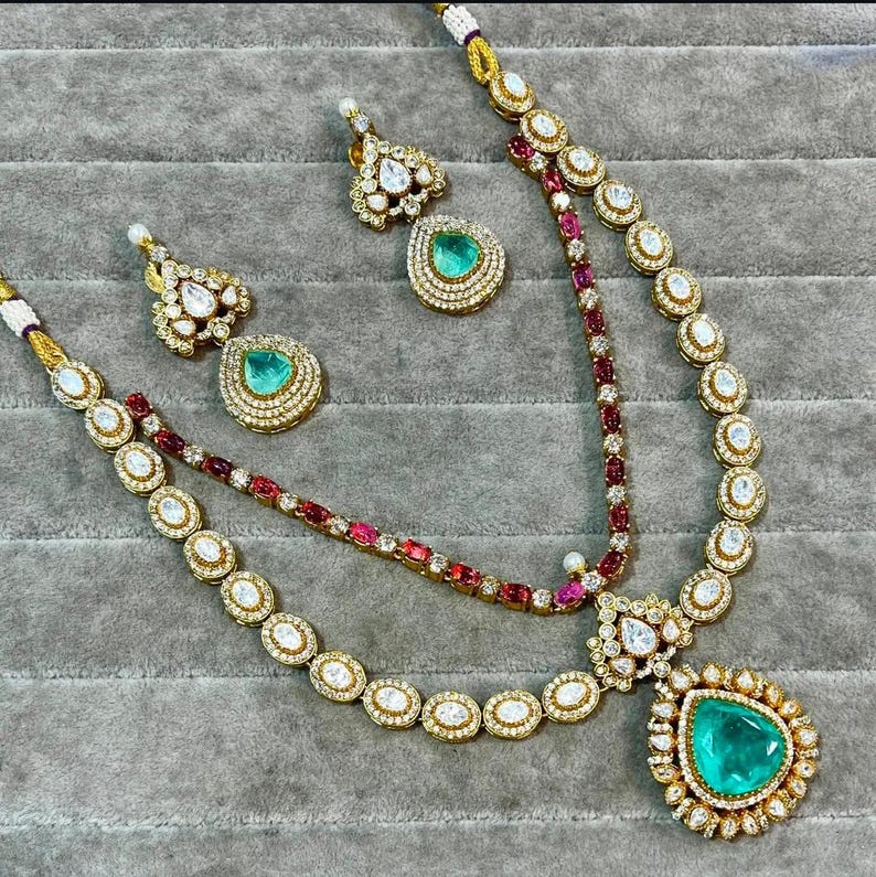 K&ouml;nnte beinhalten: Ein goldfarbenes Halsketten- und Ohrring-Set. Die Halskette hat einen gro&szlig;en, smaragdgr&uuml;nen Anh&auml;nger und kleinere Edelsteine. Die Ohrringe haben ein &auml;hnliches Design mit tropfenf&ouml;rmigen gr&uuml;nen Edelsteinen. Das Set wird auf einer grauen Oberfl&auml;che pr&auml;sentiert.