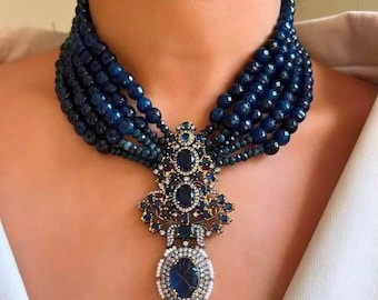 May include: A multi-strand necklace featuring dark blue beads and a detailed pendant. The pendant has an oval sapphire, surrounded by white stones and intricate gold filigree. The necklace is a statement piece.