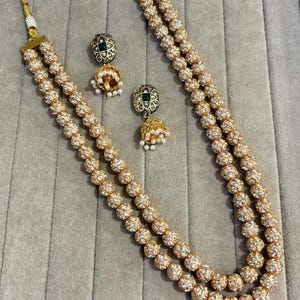 Puede incluir: Juego de collar y pendientes dorados. El collar presenta tres hilos de esferas con cuentas. Los pendientes tienen un diseño floral con un centro de piedra verde y una forma de campana colgante con perlas. Las joyas se exhiben sobre un fondo neutro.
