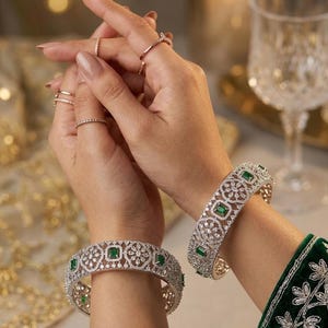 May include: Two silver-toned metal bangles featuring an intricate filigree pattern encrusted with small clear stones and rectangular green gemstones. The bangles are worn on the wrists, accompanied by several thin silver-toned rings on the fingers.