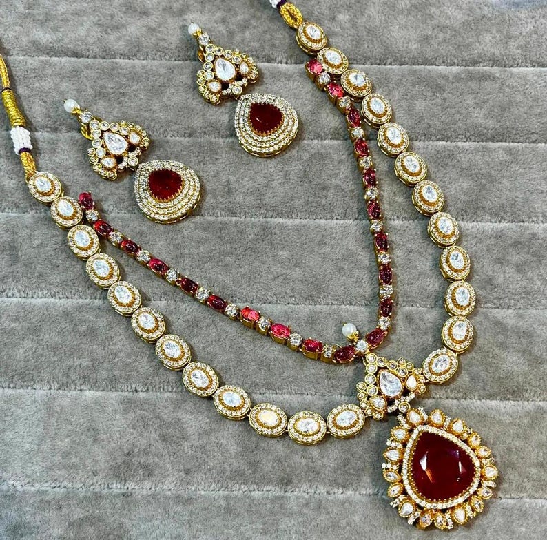 K&ouml;nnte beinhalten: Ein goldfarbenes Schmuckset mit Halskette und passenden Ohrringen. Die Halskette hat einen gro&szlig;en, tropfenf&ouml;rmigen roten Steinanh&auml;nger. Die Ohrringe und die Halskette sind mit klaren und roten Steinen besetzt.