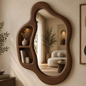 Op de afbeelding: Een wandspiegel met een onregelmatig, organisch houten frame met een ingebouwde plank met twee niveaus aan de linkerkant. De bovenste plank bevat een klein vaasje met droogbloemen, terwijl de onderste plank een boek en een klein object bevat.