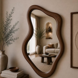 Può includere: Uno specchio da parete con una cornice irregolare dalla forma organica e ondulata, realizzata in legno marrone scuro. La superficie riflettente mostra l'interno di una stanza con un divano, un tavolino rotondo in legno e una nicchia ad arco.