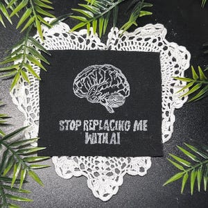 May include: A square black fabric patch featuring a white embroidered anatomical brain graphic above the text "STOP REPLACING ME WITH AI" in a bold, sans-serif font. The patch is centered on a white crocheted doily with green foliage accents.