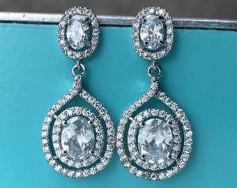 May include: A pair of silver dangle earrings with two oval-shaped clear cubic zirconia stones. The stones are set in a halo of smaller cubic zirconia stones.