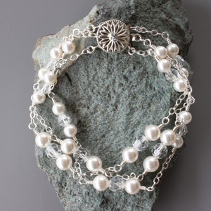 May include: A silver chain bracelet featuring white pearls and clear crystal beads. The bracelet has a silver clasp with a floral design.